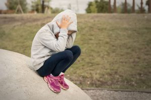 A child sits on a rock while wearing a hoodie and holding their head. This could represent the pain that depression treatment in Arcadia, AZ can address. Learn more about our services by contacting a child therapist in Phoenix, AZ or searching for child counseling Phoenix, AZ today.