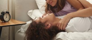 A couple smile in bed with one another next to an alarm clock. Learn how counseling Phoenix AZ can benefit your relationship with couples therapy in Phoenix, AZ. Learn more from a relationship coach in Phoenix by searching therapy in Arcadia, AZ.